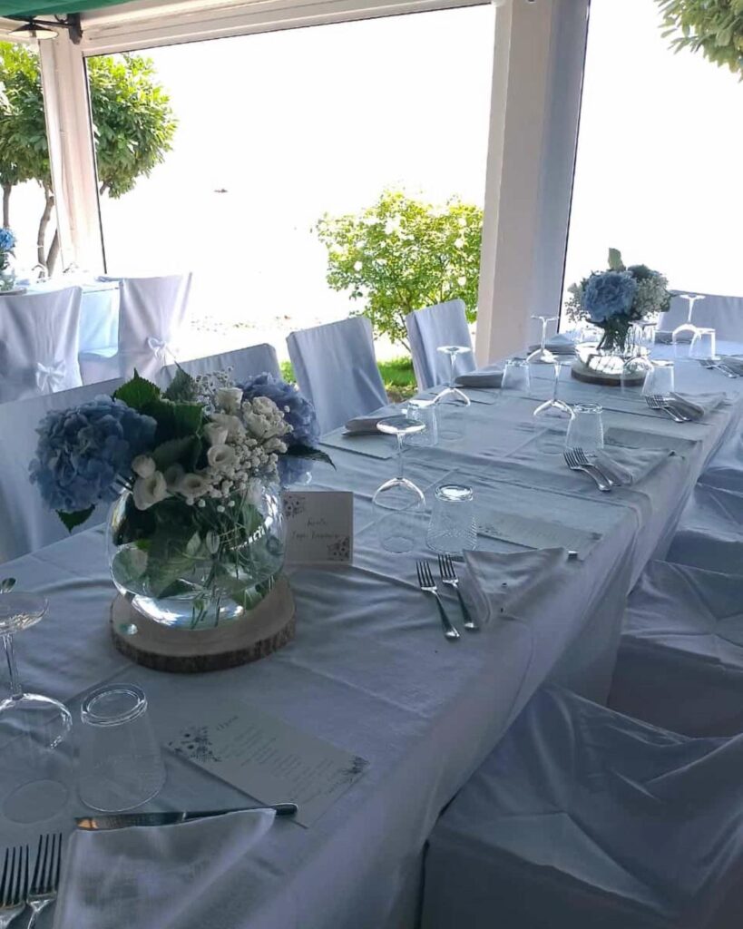 Tavola apparecchiata con tovaglia bianca e fiori blu.