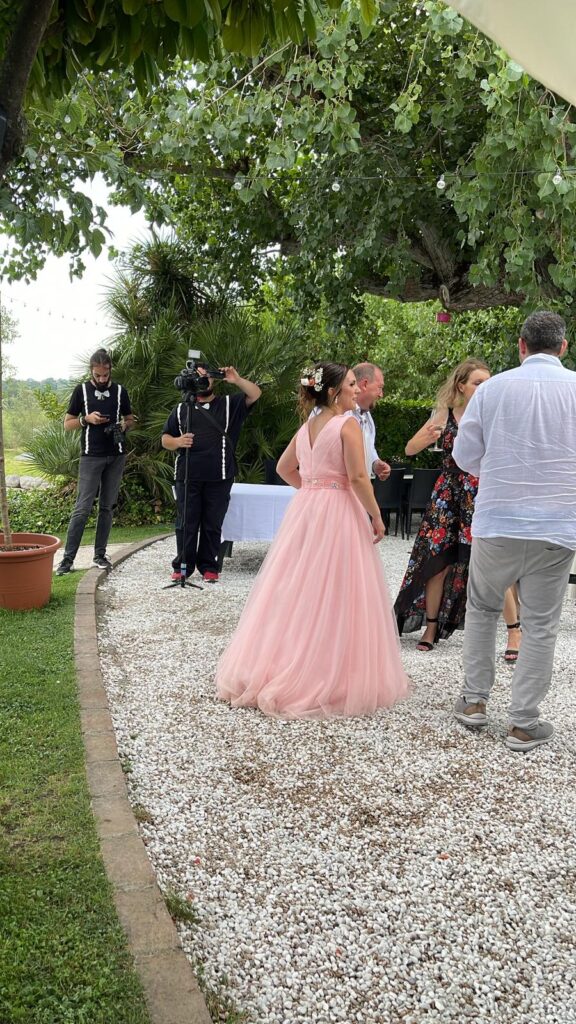 Persone in giardino, abito rosa, fotografo, alberi sullo sfondo.