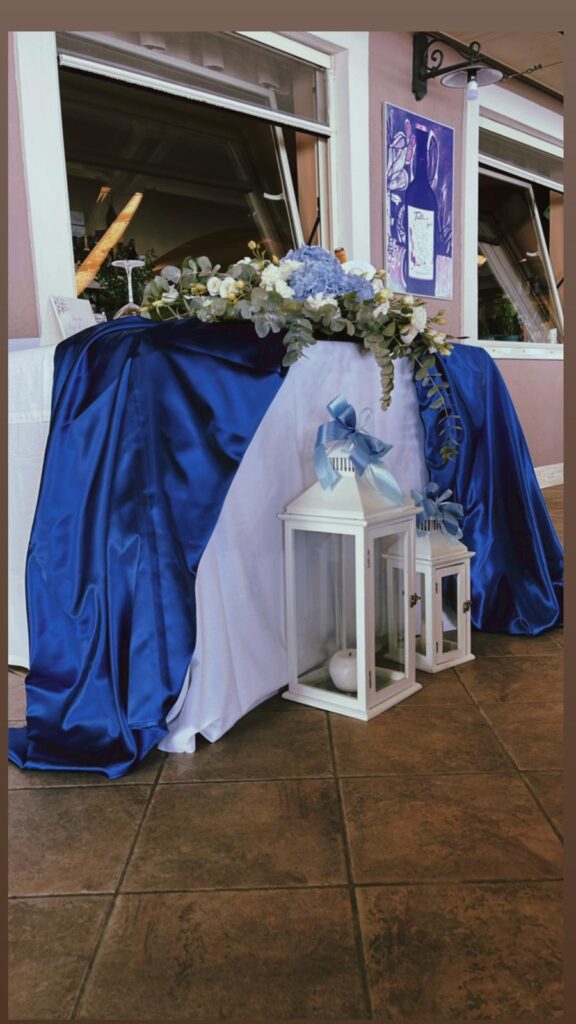 Tavolo decorato con fiori, stoffa blu e lanterne bianche.